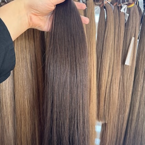 Puede incluir: Una mano sostiene un mechón de extensiones de cabello castaño oscuro. Varios otros mechones de extensiones de cabello en varios tonos de marrón y rubio son visibles en el fondo, colgados de un estante.