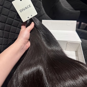 Puede incluir: Mechón de extensiones de cabello oscuro y brillante. Una etiqueta blanca con la palabra "DIVAGE" está adjunta. El cabello se sostiene en una mano, con una caja blanca visible en el fondo. El cabello es de un color negro intenso.