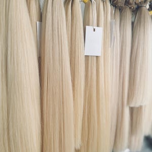 Puede incluir: Extensiones de cabello rubio colgadas de una barra de metal. Las extensiones están agrupadas y atadas con pequeñas bandas. Una etiqueta blanca con la palabra "DIVAGE" es visible. El cabello tiene una textura suave y lisa y un color rubio claro.