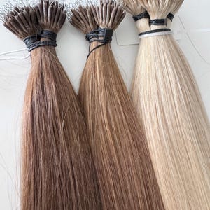 以下が含まれることがあります： 茶色とブロンドのさまざまな色合いのヘアエクステンションの3つの束。各束は、黒いバンドと上部の金属ループで固定されています。髪はストレートで滑らかに見え、自然な光沢があります。