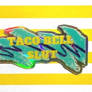 May include: A holographic sticker with the text "TACO BELL SLUT" in yellow block letters. The sticker has a teal background with purple and blue accents, and a red and yellow border. The sticker is on a yellow and white striped background.