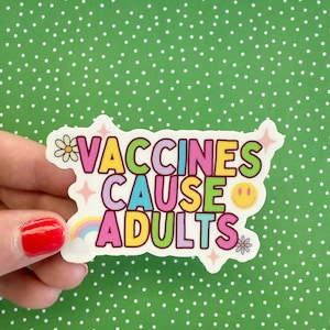 May include: A colorful sticker with the text "VACCINES CAUSE ADULTS" in various colors. The sticker includes a rainbow, a smiley face, and flower graphics. The background is a green surface with white polka dots.