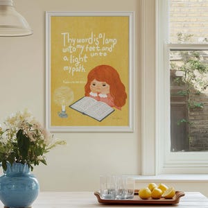 May include: A framed print with a yellow background features an illustration of a girl with red hair reading a book. The text "Thy word is a lamp unto my feet, and a light unto my path" is written above the girl. A blue vase with white flowers sits on a wooden table.