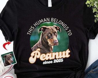 May include: Black t-shirt with a circular graphic featuring a brown dog and the text "THIS HUMAN BELONGS TO Peanut since 2025". The graphic is surrounded by a green background.