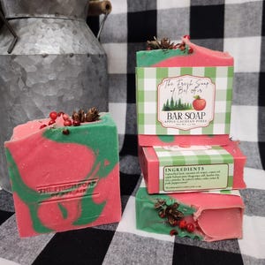 May include: A stack of bar soaps with a red and green swirl design, topped with berries and pine cones. The soap is labeled "The Fresh Soap of Bel Air" and "Apple-Lachian Pines". The background features a black and white checkered pattern.