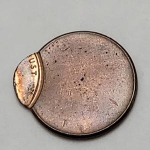 May include: A copper-colored coin with a gold-colored attachment. The attachment has the letters "UST" printed on it. The coin has a circular shape with a slightly irregular edge.
