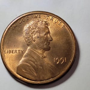 May include: A copper-colored United States one-cent coin featuring the profile of Abraham Lincoln facing right. The text IN GOD WE TRUST is arched across the top, the word LIBERTY is on the left, and the year 1991 is printed on the right side.