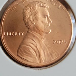May include: A copper-colored United States penny coin. The obverse features a profile of Abraham Lincoln, the word "LIBERTY", and the year 2025. The coin also has the inscription "IN GOD WE TRUST".