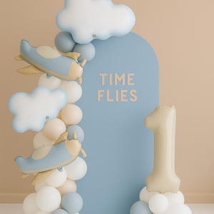 May include: A blue arched backdrop with the text TIME FLIES in beige letters, decorated with a balloon garland featuring white, beige, and blue balloons, two airplane-shaped foil balloons, two cloud-shaped foil balloons, and a large number 1 balloon.