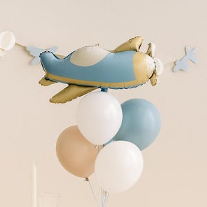 May include: A festive scene with a blue and gold airplane balloon, white, blue, and tan balloons, paper airplanes, and cookies. The table is covered with a light-colored checkered tablecloth, creating a whimsical party atmosphere.