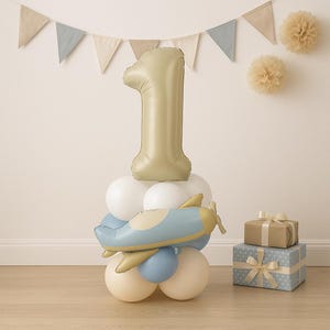 May include: A festive first birthday decoration featuring a large gold number one balloon atop a cluster of white, blue, and beige balloons. A blue and yellow airplane balloon is also included. Gift boxes and a banner complete the celebratory scene.