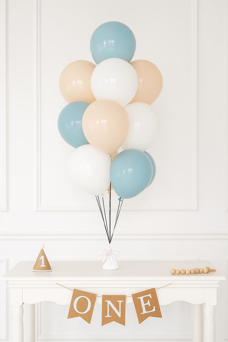 May include: A cluster of pastel-colored balloons, including blue, white, and beige, tied together. Below, a white table displays a banner that spells "ONE", a party hat, and wooden beads. The balloons are a festive decoration.