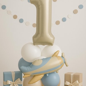 May include: A festive arrangement for a first birthday celebration. A large gold number one balloon sits atop a cluster of white and blue balloons, with a toy airplane. Several wrapped gifts in blue and brown with ribbons complete the scene.