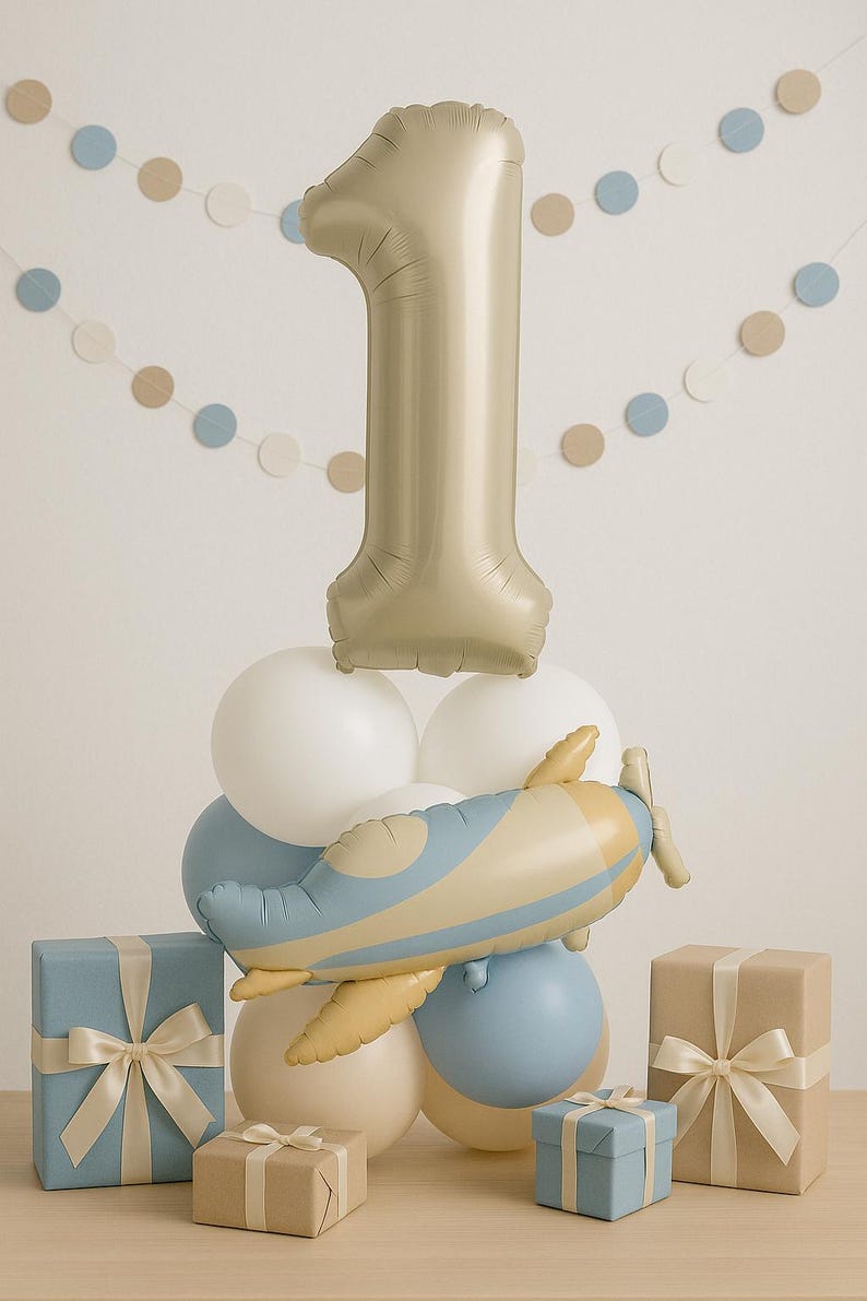 May include: A festive arrangement for a first birthday celebration. A large gold number one balloon sits atop a cluster of white and blue balloons, with a toy airplane. Several wrapped gifts in blue and brown with ribbons complete the scene.