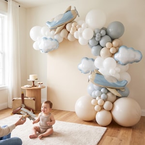 May include: A balloon arch featuring white, beige, and grey balloons, accented with two light blue airplane-shaped foil balloons and two cloud-shaped foil balloons. The arrangement is displayed as a decorative backdrop for a celebration.