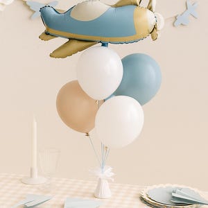 May include: A balloon bouquet featuring a large foil airplane balloon in dusty blue and gold, accompanied by four round latex balloons in white, dusty blue, and beige. The balloons are anchored to a small white base with thin blue ribbons.