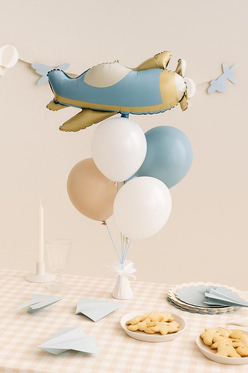 May include: A balloon bouquet featuring a large foil airplane balloon in dusty blue and gold, accompanied by four round latex balloons in white, dusty blue, and beige. The balloons are anchored to a small white base with thin blue ribbons.