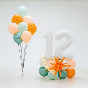 May include: A festive birthday arrangement featuring a cluster of colorful balloons in shades of orange, peach, teal, and mint green. Beside it, a display of white number balloons spelling out "12", accented with orange starburst decor and additional balloons.