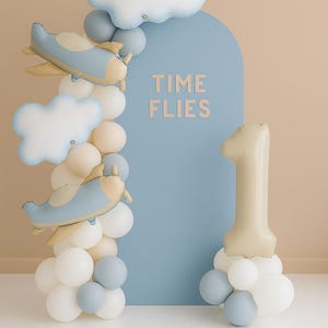 May include: A festive balloon arrangement for a first birthday celebration. Features a blue arch with the text "TIME FLIES", airplane and cloud balloons, and a large number one balloon. Balloons are in shades of blue, white, and beige.