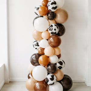 May include: A balloon garland in a vertical arrangement, featuring white, brown, tan, and cow-print balloons. The garland is designed for party decorations, with a mix of sizes and shades of brown.
