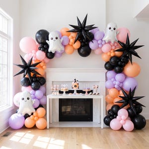 Peut inclure: Une arche de ballons d'Halloween composée de ballons noirs, orange, violet clair et roses. L'ensemble comprend des ballons fantômes en aluminium, des étoiles noires et une table centrale avec un gâteau portant le chiffre 1.