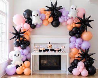 May include: A Halloween balloon arch featuring black, orange, light purple, and pink balloons. The display includes foil ghost balloons, black starburst balloons, and a central table with a cake topper displaying the number 1.