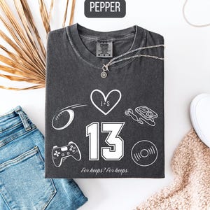 May include: A dark gray t-shirt with white graphics including a heart, football, game controller, and a music disc. The number 13 is prominently displayed. Text at the bottom reads "For keeps? For keeps."