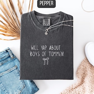May include: A dark gray t-shirt with the text "WILL YAP ABOUT BOYS OF TOMMEN" in white. The shirt is paired with blue jeans, a silver necklace, and white slip-on shoes. A dried palm leaf and a beige blanket are also in the image.