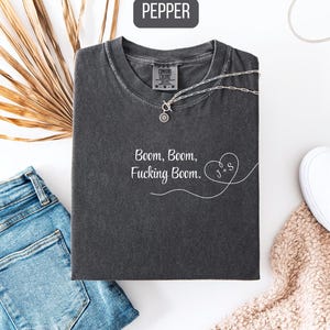 May include: A dark gray t-shirt with the text "Boom, Boom, Fucking Boom" in white script. The shirt is paired with blue jeans, white sneakers, and a silver necklace. The word "Pepper" is in a black box in the top left corner.