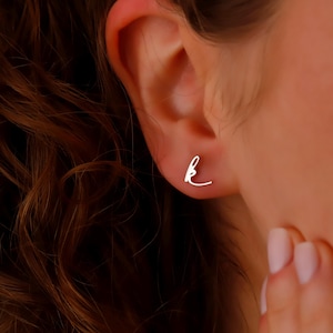 May include: A pair of small, silver-colored stud earrings in the shape of the cursive letter "k" are shown. The earrings are worn on an ear, with the person's hair and part of their face visible in the background. The earrings are simple and elegant.