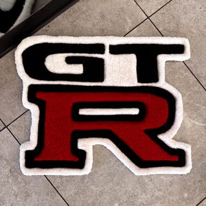 Puede incluir: Una alfombra personalizada con las letras "GT-R" en un diseño llamativo. Las letras son negras y rojas, contorneadas en negro, sobre un fondo blanco. La alfombra está colocada sobre un suelo embaldosado.