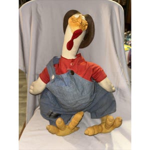 Puede incluir: Una muñeca de peluche de pavo con una camisa roja y un peto vaquero. El pavo tiene un sombrero marrón, un cuello largo blanco y patas amarillas. La muñeca está sentada sobre una superficie blanca.