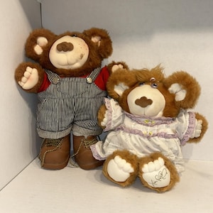 Puede incluir: Dos osos de peluche. Uno lleva una camisa roja, un peto a rayas y botas marrones. El otro oso lleva un vestido blanco con ribete de encaje morado. Ambos osos tienen pelaje marrón y caras amigables.