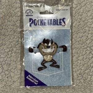 May include: A Looney Tunes Pocketable featuring Taz, the Tasmanian Devil, in a plastic package. The Pocketable is brown, tan, and black, designed to look like it's popping out of a pocket. The packaging reads "Pocketables" and "Makes a Great Bookmark Too!"