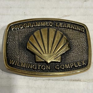 以下が含まれることがあります： 「PROGRAMMED LEARNING」と「WILMINGTON COMPLEX」の文字が刻まれた、長方形の真鍮色のベルトバックル。バックルの中央には、スタイリッシュな貝殻のデザインがあり、背景はテクスチャ加工されています。