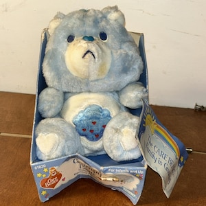 May include: A light blue plush bear toy with a white belly patch featuring a blue cloud and red raindrops. The bear has dark blue eyes and a blue nose. It is housed in a blue cardboard box with the text The CARE BEARS Family is Growing.