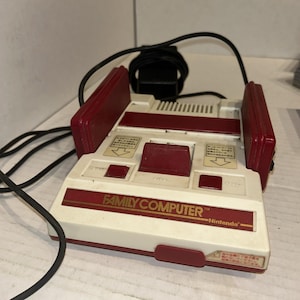 Può includere: Una console per videogiochi Nintendo Family Computer vintage. La console è bianca con dettagli bordeaux e presenta la scritta "FAMILY COMPUTER" e il logo Nintendo. Ha un cavo di alimentazione e porte per controller.