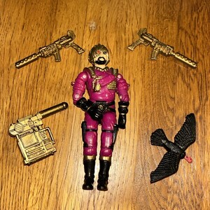 Peut inclure: Une figurine d'action vintage en uniforme magenta avec des bottes et des gants noirs, entourée d'armes dorées et d'un oiseau noir. La figurine a une barbe et un œil rouge. Les accessoires comprennent deux fusils, un pistolet et un oiseau.