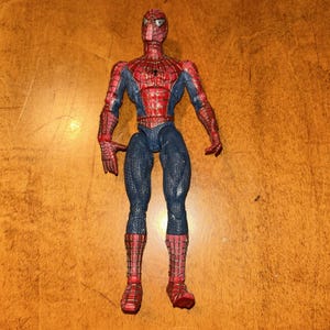 May include: A Spider-Man action figure featuring a red and blue suit with a black web pattern across the chest, arms, and legs. The figure has white triangular eyes, a small black spider emblem on the chest, and a textured surface throughout.