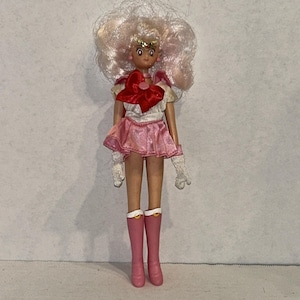 Könnte beinhalten: Puppe mit hellrosa Haaren und einem rosa-weißen Outfit. Die Puppe hat eine rote Schleife auf der Brust, einen rosa Rock und rosa Stiefel. Die Puppe trägt weiße Handschuhe und eine goldene Tiara.