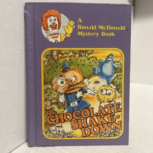 Könnte beinhalten: Ein lila Buch mit dem Titel "A Ronald McDonald Mystery Book" mit Illustrationen. Das Cover zeigt Ronald McDonald, einen Burger-Charakter mit einer Lupe und dem Titel "The Chocolate Shake-Down".