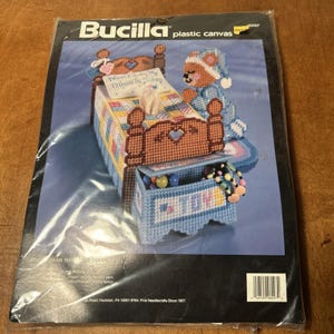 May include: A Bucilla plastic canvas craft kit package featuring a finished tissue box cover shaped like a bed with a teddy bear, a quilt, and a toy chest. The text on the pillow reads Now I Lay Me Down To Sleep and the toy chest is labeled TOYS.