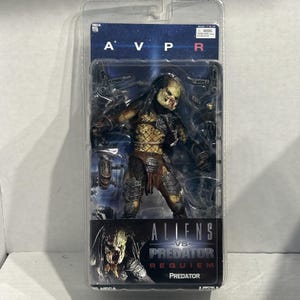 Puede incluir: Una figura de acción de Predator empaquetada de la película "Aliens vs. Predator: Requiem". La figura está en una caja de plástico transparente. El Predator es una figura detallada y multicolor con cabeza amarilla, armadura marrón y pose amenazante.