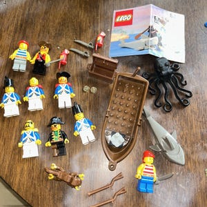 May include: A collection of LEGO pirate-themed minifigures, including soldiers in blue uniforms and pirates, a brown plastic boat, a black octopus, a gray shark, two parrots, a treasure chest, two gold coins, two brown oars, and a LEGO instruction manual.