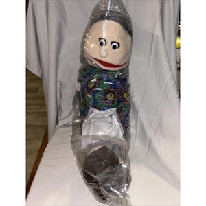 May include: A vintage puppet with a fabric face, black hair, and a red mouth. The puppet is wearing a blue and green floral patterned dress and a white apron. The puppet is enclosed in a clear plastic bag.