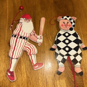 Op de afbeelding: Twee houten kerstversieringen. Eén is de kerstman in een honkbaluniform, met een knuppel. De andere is een varken in een zwart-wit ruitpatroon outfit. Beide hebben een rood touwtje om op te hangen.