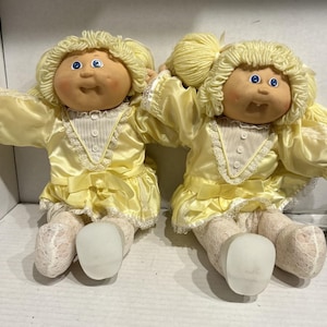 May include: Two dolls with light skin, blue eyes, and pale yellow yarn hair. They wear matching pale yellow satin dresses featuring white lace trim, a vertical striped bodice with buttons, and a bow at the waist. They have white lace leggings.