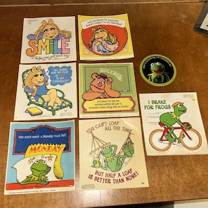 Può includere: Una collezione di adesivi vintage dei Muppet con Kermit la Rana, Miss Piggy e Fozzie. Gli adesivi presentano varie citazioni e frasi, tra cui "Smile", "I Brake for Frogs" e "But half a loaf is better than none!"