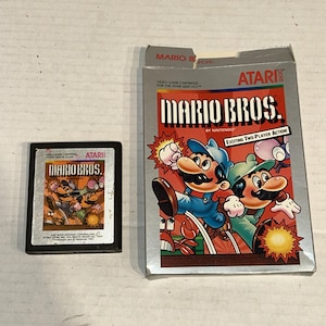 Peut inclure: Une cartouche et une boîte de jeu vidéo Atari Mario Bros. La boîte présente Mario et Luigi avec le texte "Mario Bros." et "Exciting Two-Player Action !" La cartouche a un design similaire.