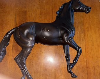 May include: A dark brown horse figurine in a trotting pose. The horse has a detailed mane and tail, with some visible wear and tear on the surface. The figurine is made of a solid material and is displayed on a wooden surface.